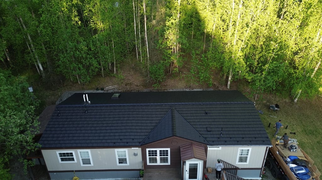A metal roof on a ranch-style home with a ridge cap, roof louver, and vent pipe, surrounded by lush greenery in a rural forested area.