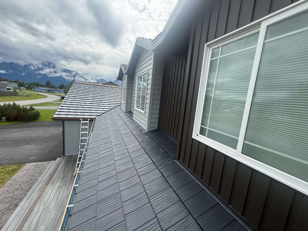 A modern home with a dark grey metal roof, set against a lush green mountain backdrop under an overcast sky.