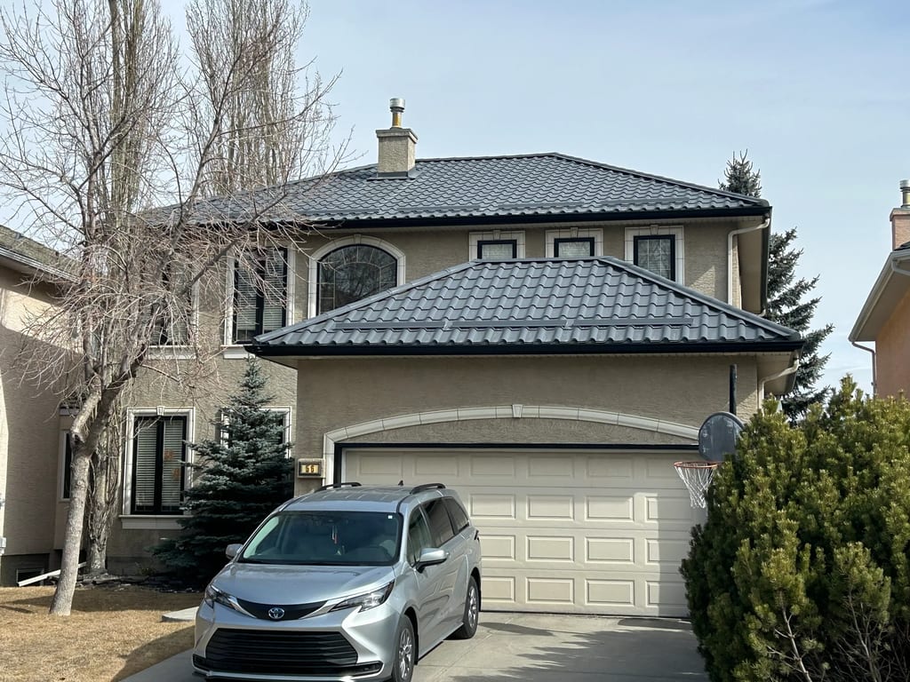 Charcoal Grey Interlock Tile aluminum roof on a beige two-storey home with dormers among tall pines, Calgary, Alberta