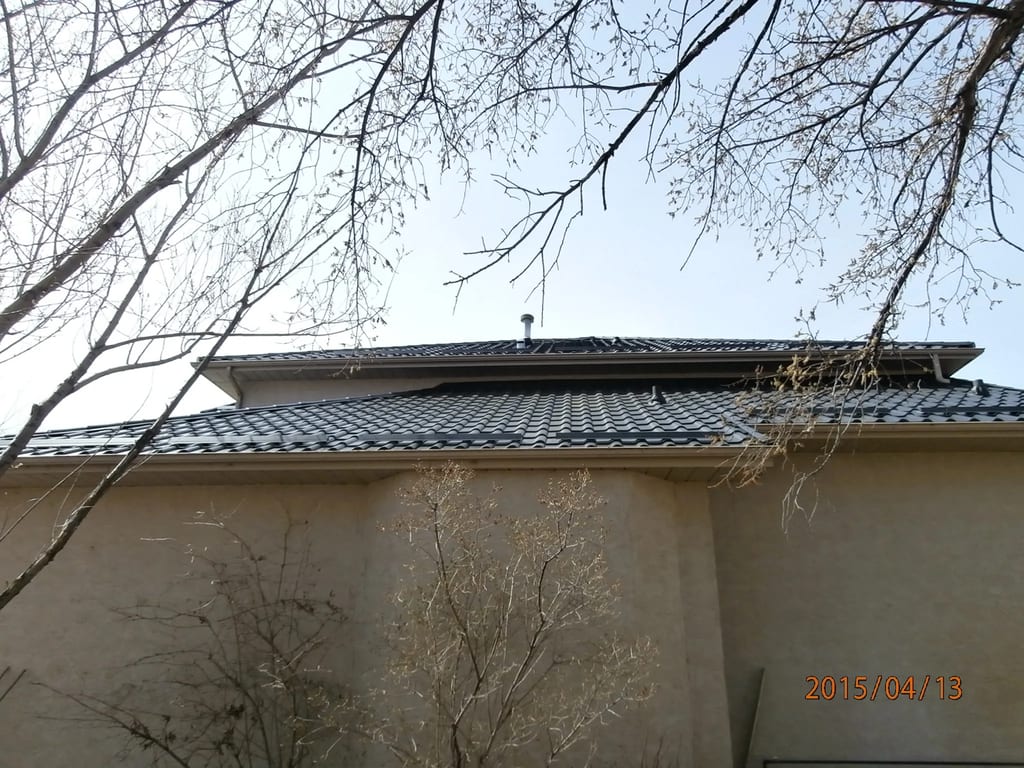 Black Interlock Tile aluminum roof on a beige two-storey home with dormers among bare trees, Redcliff, Alberta
