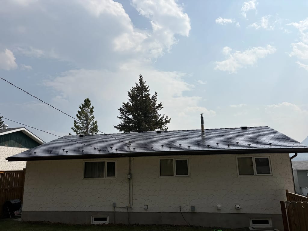Deep Charcoal Interlock Slate aluminum roof on a white ranch-style home with a metal roof among tall trees, Coleman, Alberta