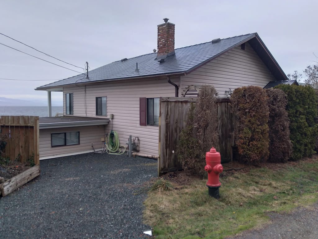 Black Interlock Slate aluminum roof on a white home with a chimney among tall bushes and trees, Qualicum, British Columbia