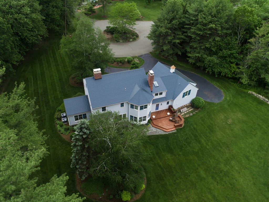 Aerial rear view of a large Colonial home in Topsfield, MA with a complex deep charcoal Interlock aluminum slate metal roof, two chimneys, and a rear wood deck.