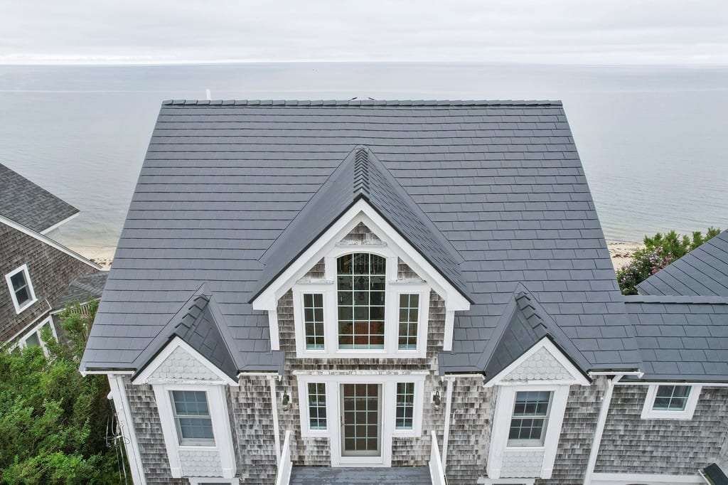 Aerial front view of a Cape Cod beach house with a complex deep charcoal Interlock aluminum slate metal roof featuring multiple dormers and valleys, with ocean backdrop.