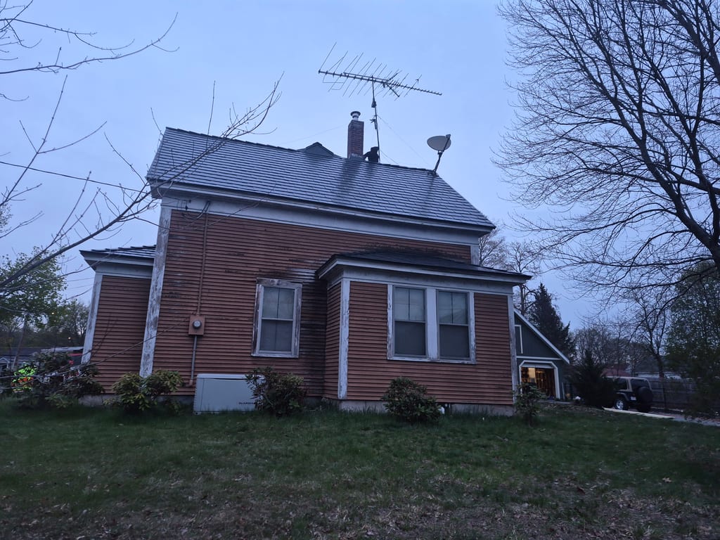 Heritage-style single-family home in Ayer, MA with deep charcoal Interlock aluminum slate mansard roof, brick chimney, and satellite dish at dusk.