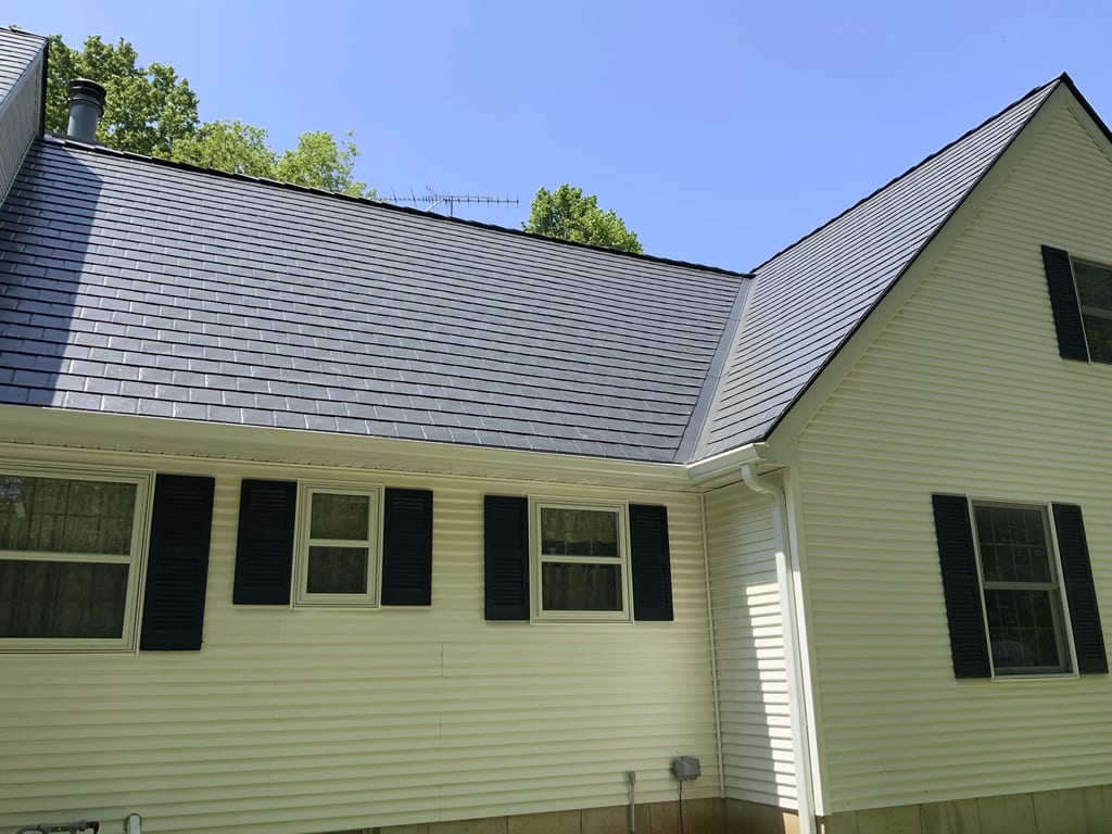Rear-left view of a Colonial home with Interlock black aluminum slate metal roofing, white vinyl siding, and black shutters under a clear blue sky.