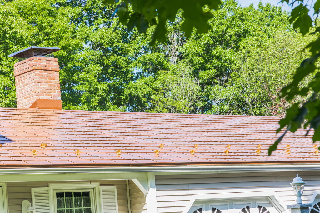 Ranch-style home with Interlock aged-copper aluminum slate metal roofing, brick chimney with cap, and lush green tree backdrop in North Hampton, NH.