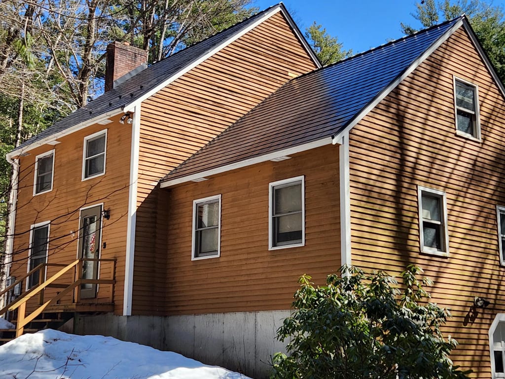 Two-story colonial home with black Interlock aluminum slate metal roofing, warm cedar lap siding, and a brick chimney set in a forested New Hampshire winter landscape.