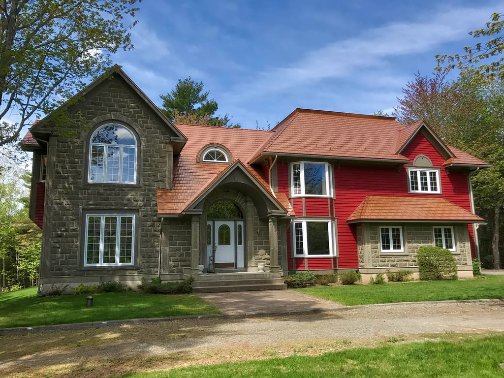 Black Interlock Slate aluminum roof on a red two-storey home with dormers among tall pines, Reids Hill (Shelburne), Nova Scotia