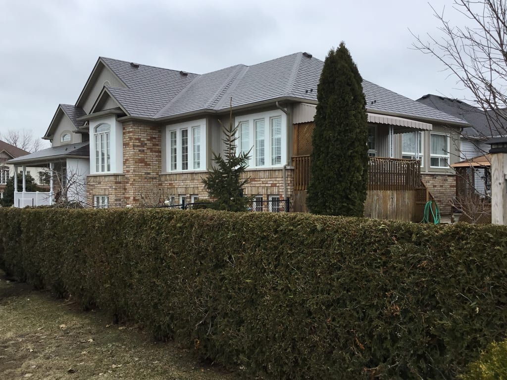 Large single-family home with complex multi-hip Interlock aluminum slate roof in Tudor Brown, brick exterior, viewed from front-right across a trimmed cedar hedge.