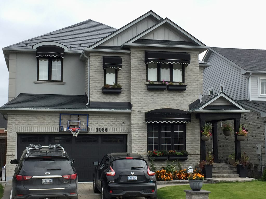 Two-storey craftsman home in Milton ON with black Interlock aluminum slate metal roofing, light grey brick facade, black awnings, and window boxes.