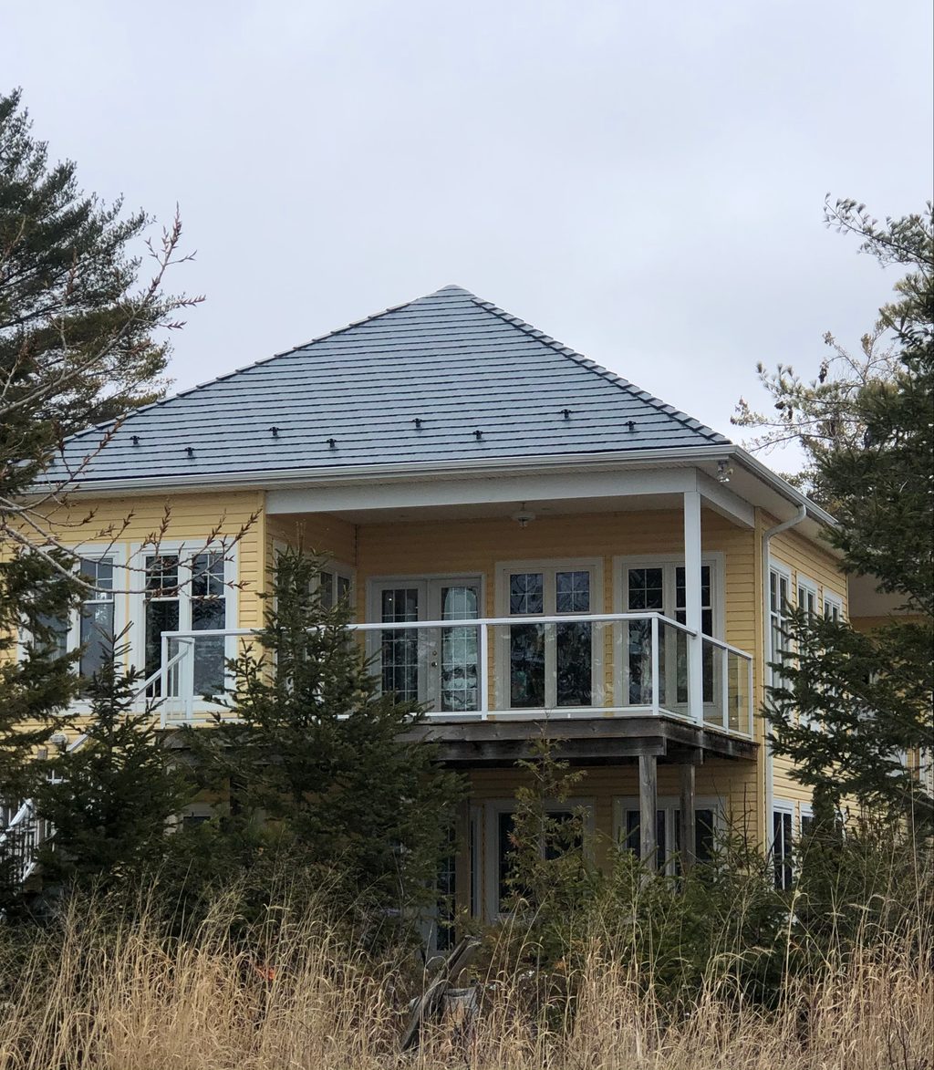 Two-storey yellow cottage with a hip-profile deep charcoal Interlock cedar shingle metal roof, snow guards, and a covered upper deck balcony, set among coastal grasses and pines.