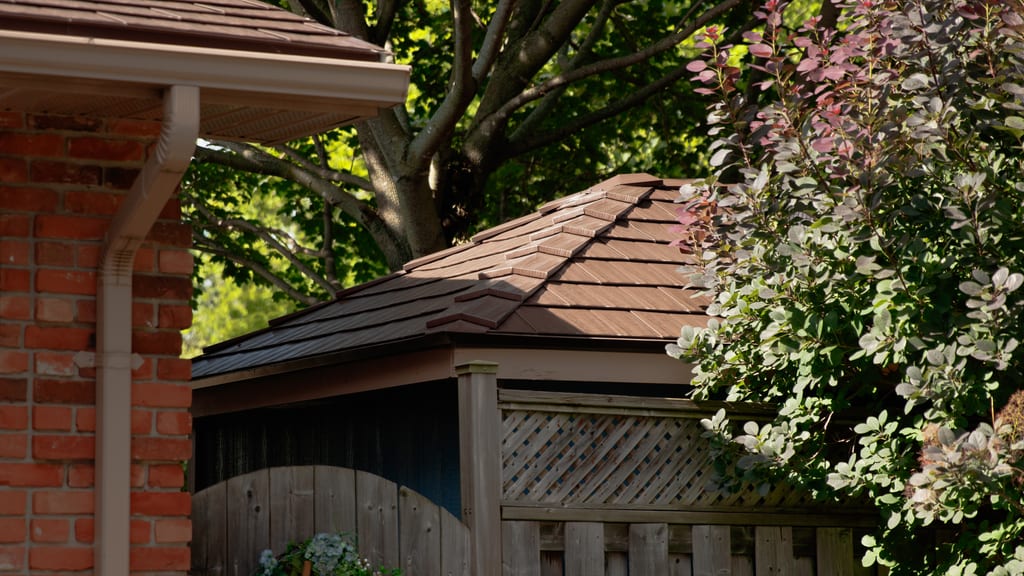 Interlock Colonial Brown cedar shingle aluminum hip roof on a small gazebo or hot tub enclosure beside a brick home, surrounded by lush garden greenery.