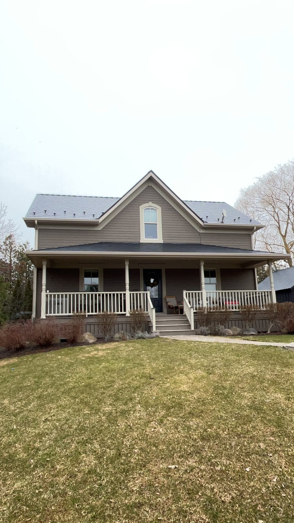Two-storey farmhouse in Ravenna, ON with black Interlock aluminum slate metal roof, front dormer, wrap-around porch, and snow guards visible on steep gable.