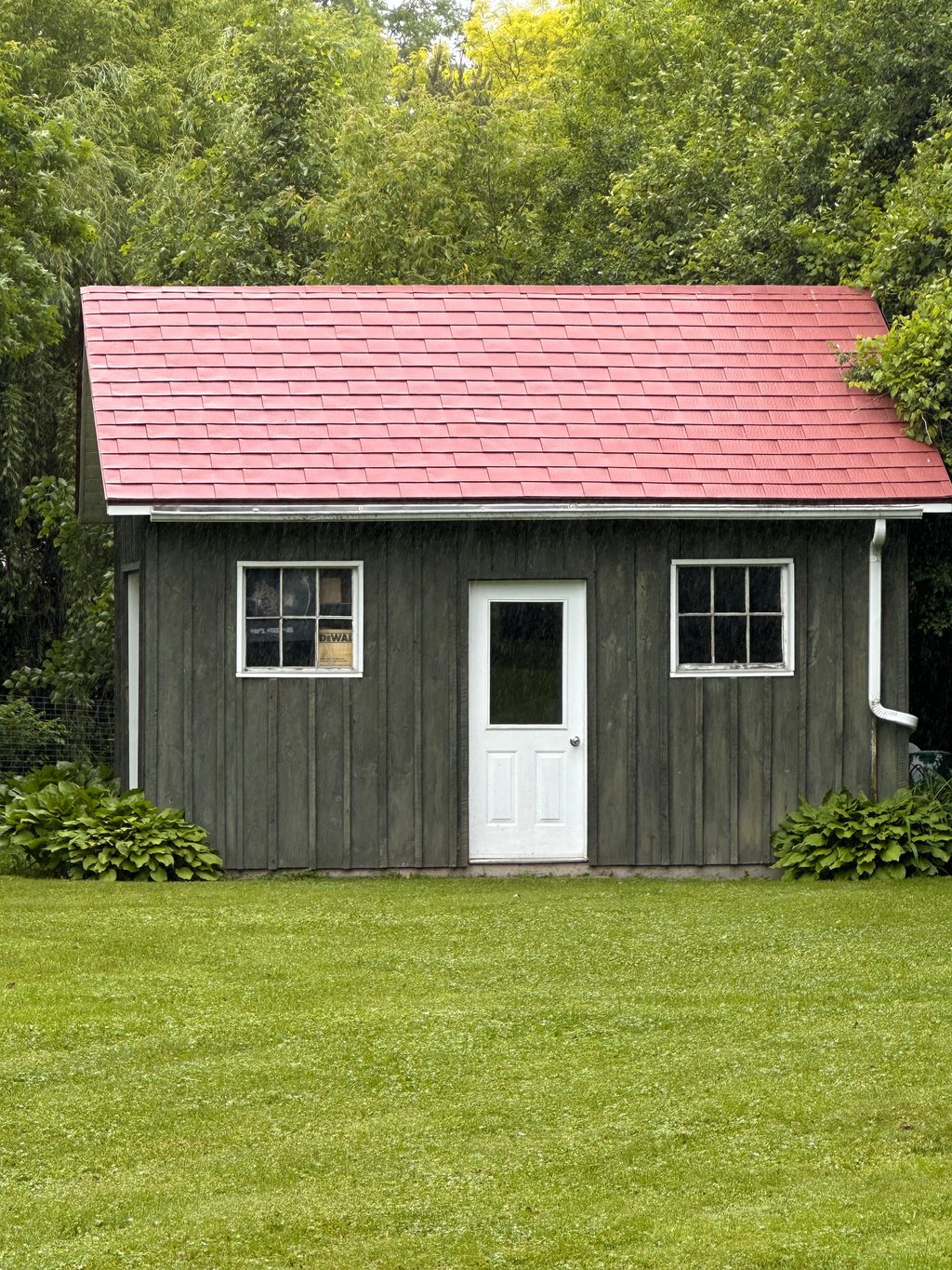 Front elevation of a rustic wood-clad outbuilding with a Tile Red Interlock aluminum shingle gable roof, white door, and twin windows in Almonte, ON.