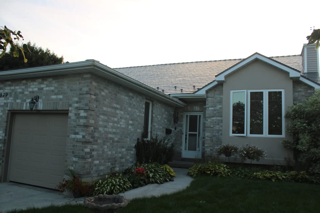 Deep Charcoal Interlock Cedar Shingle aluminum roof on a tan one-storey home with a metal roof among trees, Kingston, Ontario