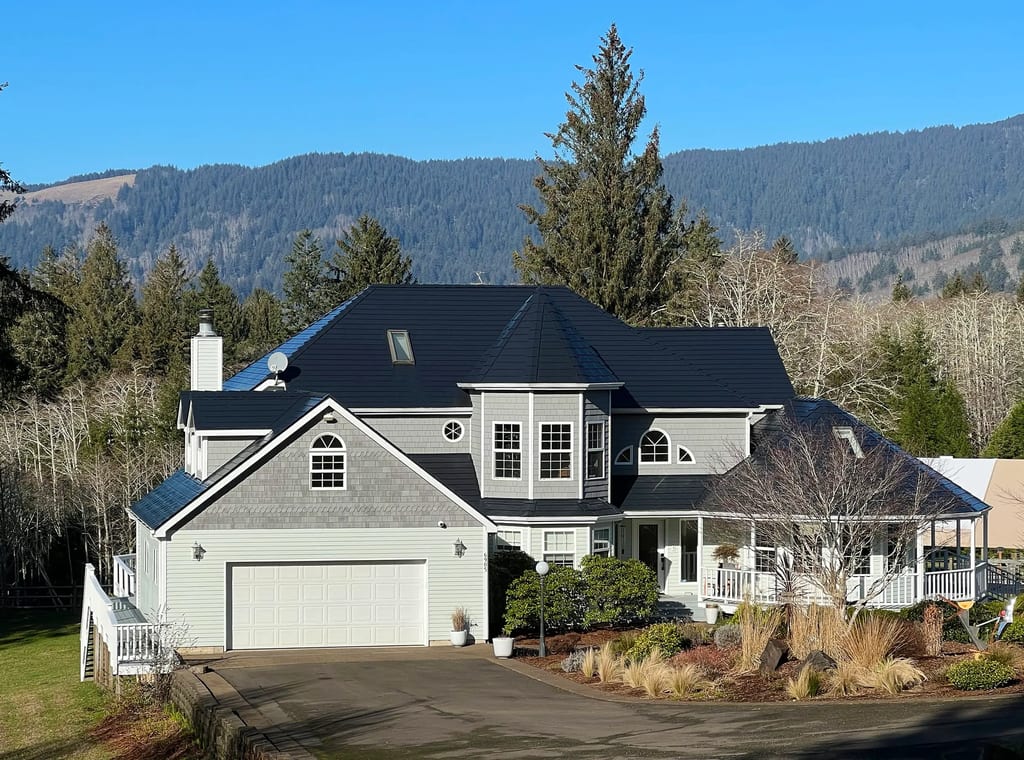 Two-story gray shingle-sided home in Otis, OR with a complex black Interlock aluminum slate metal roof, chimney, skylight, and mountain backdrop.