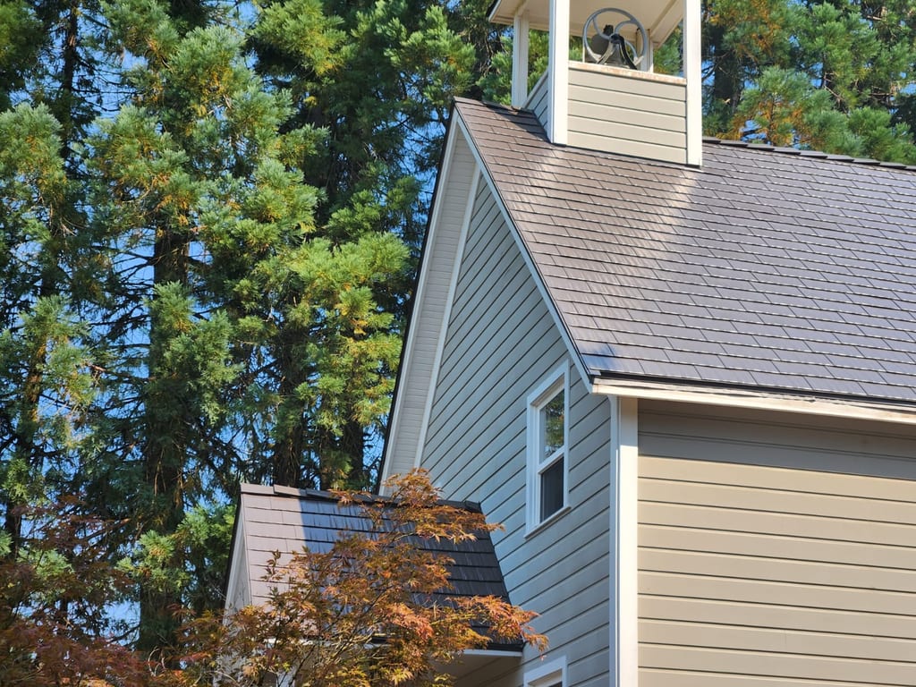 Steep-pitched Tudor Brown Interlock cedar shingle metal roof on a gray-sided church with bell tower cupola, set among tall Pacific Northwest conifers.