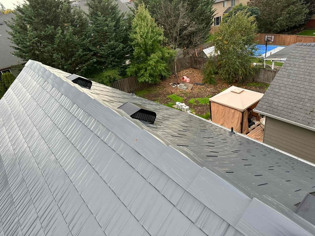 Aerial ridge-line view of a deep charcoal Interlock aluminum cedar shingle metal roof with ridge cap and roof louvers on a suburban two-story home.