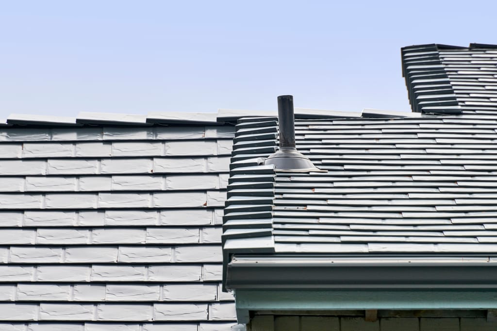 Close-up of Interlock deep charcoal aluminum slate roofing showing multi-level roof transition with vent pipe flashing detail against a clear blue sky.