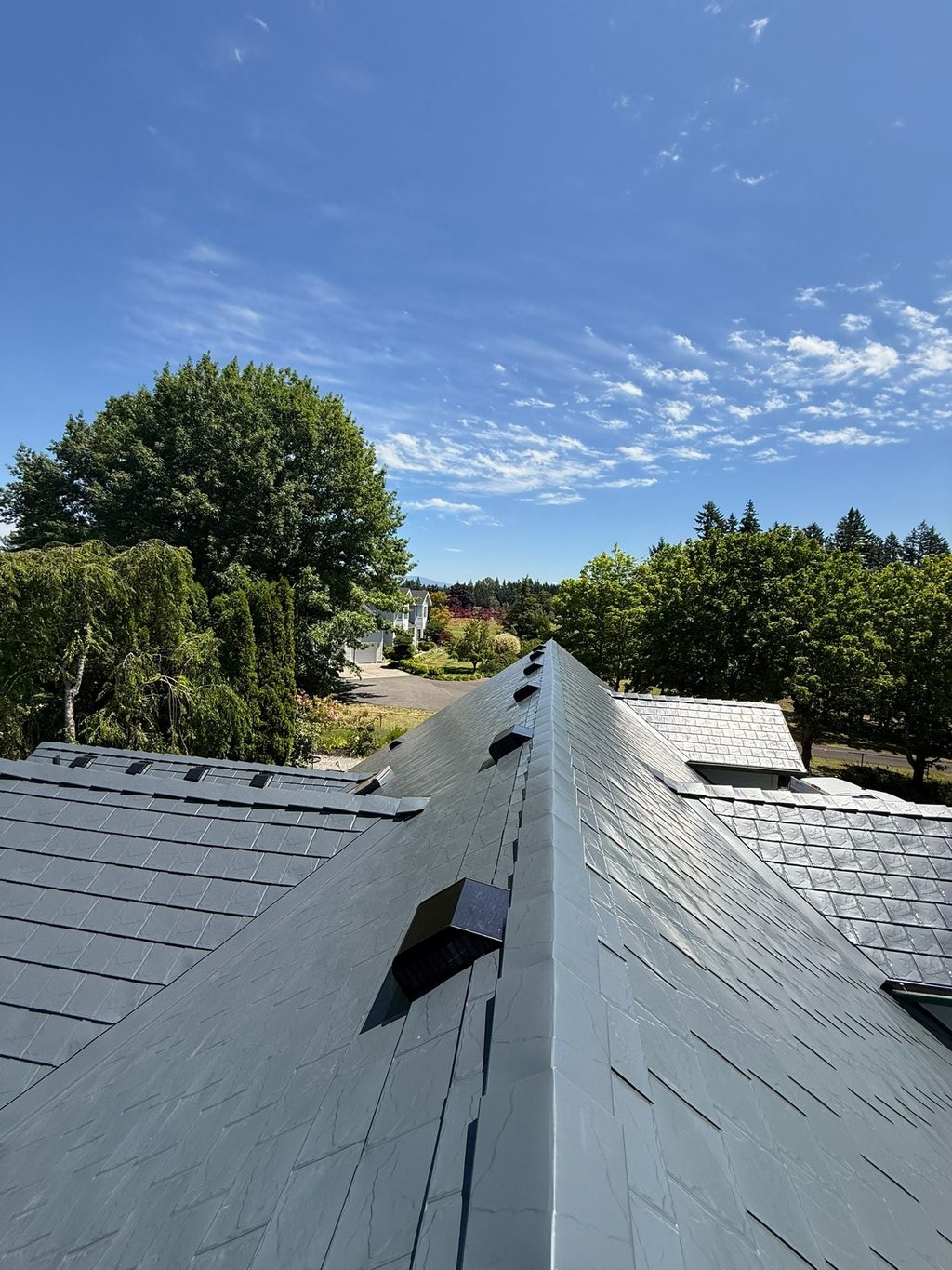 Looking down the steep ridge of a deep charcoal Interlock aluminum slate roof on a two-story suburban home, with lush trees and blue sky beyond.