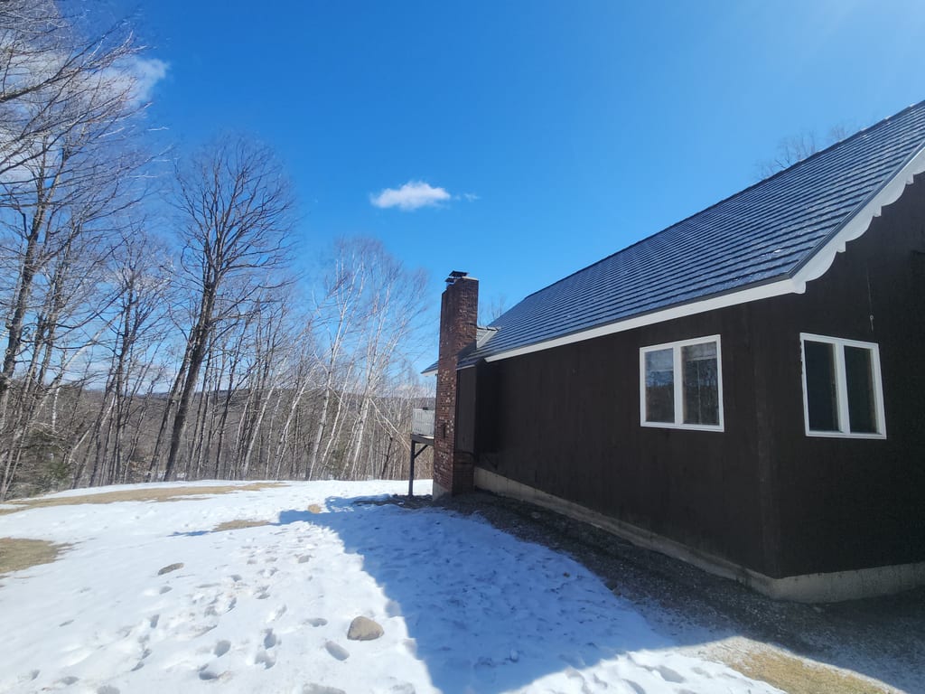 Musket Grey Interlock cedar shingle metal roof on a dark-brown ranch home in Vermont, featuring a brick chimney, surrounded by bare winter trees.