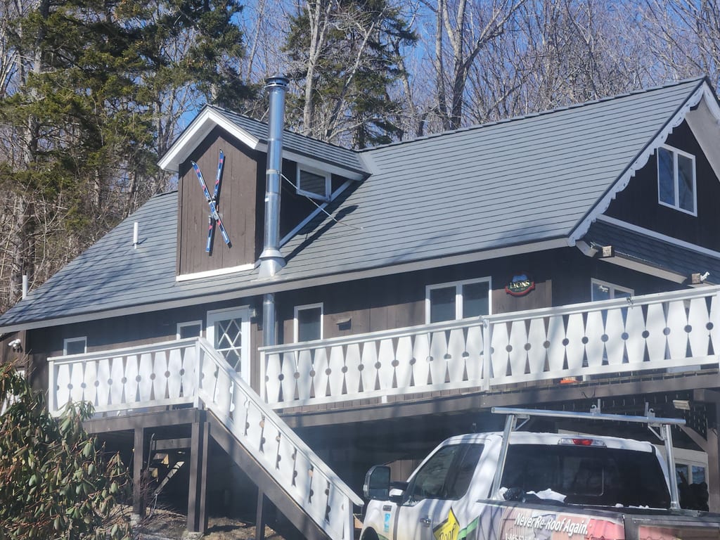 Two-story chalet-style Vermont home with Interlock Musket Grey cedar shingle aluminum roof, front dormer, vent pipe, and white deck railings amid winter forest.