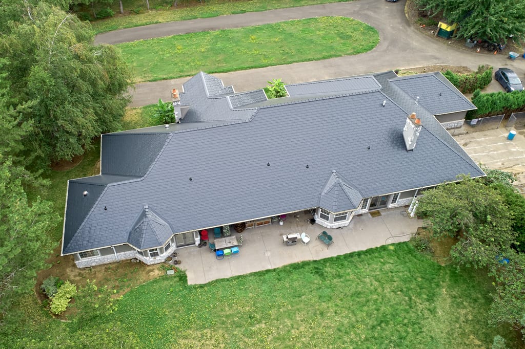 Aerial rear view of a large ranch-style home in Ridgefield, WA with a complex Interlock aluminum slate roof in charcoal black, featuring turreted dormers and two chimneys.