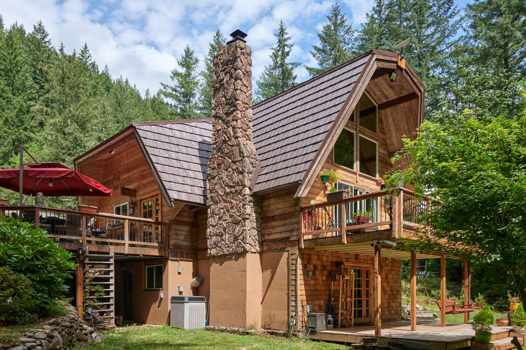 Rear-left view of a forested chalet with steep Colonial Brown Interlock aluminum shake roof, massive stone chimney, and multi-level wood decks.