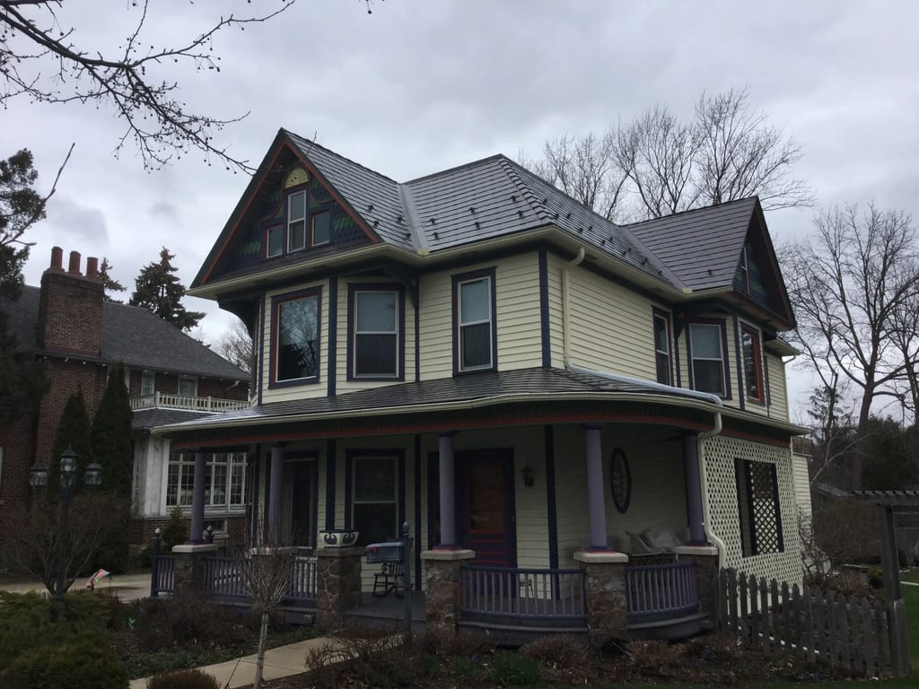 Weathered Interlock Slate aluminum roof on a cream two-storey home with dormers and a porch among tall, Mayville, Wisconsin