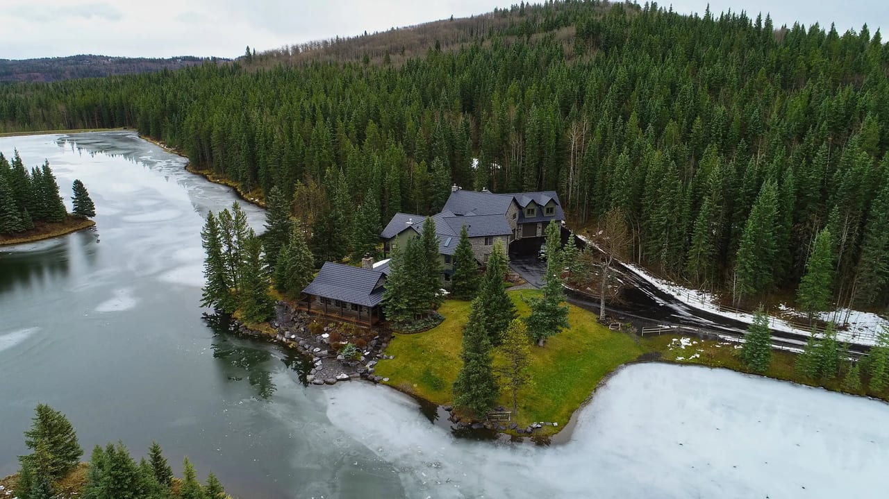 Aerial view of a chalet-style home on a lakeside peninsula in Bragg Creek AB featuring black Interlock aluminum shake metal roofing with complex multi-gable design.