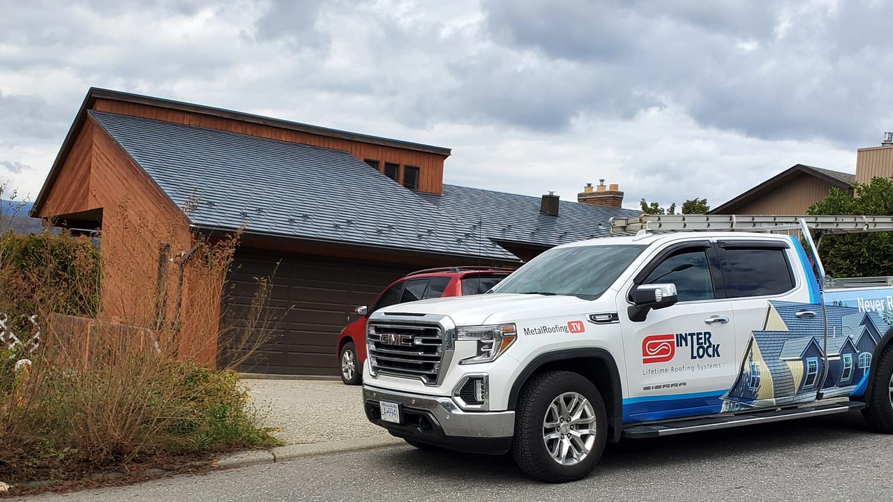 Slate-blue Interlock Steel Slate metal roof on a 1.5-storey Craftsman home in Vernon BC, viewed from the front-left with an Interlock GMC Sierra service truck in foreground.