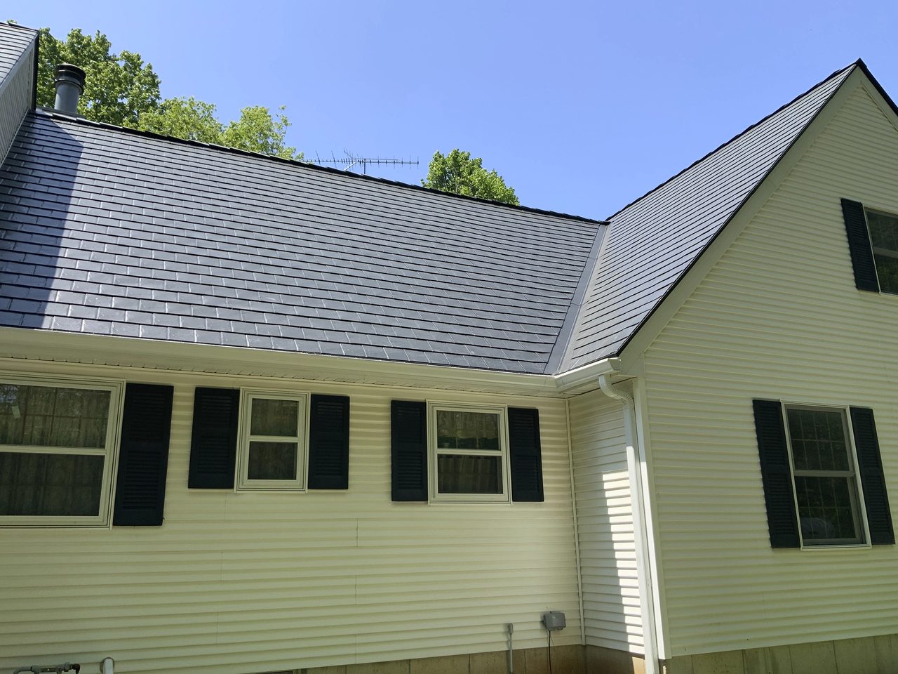 Rear-left view of a Colonial home with Interlock black aluminum slate metal roofing, white vinyl siding, and black shutters under a clear blue sky.
