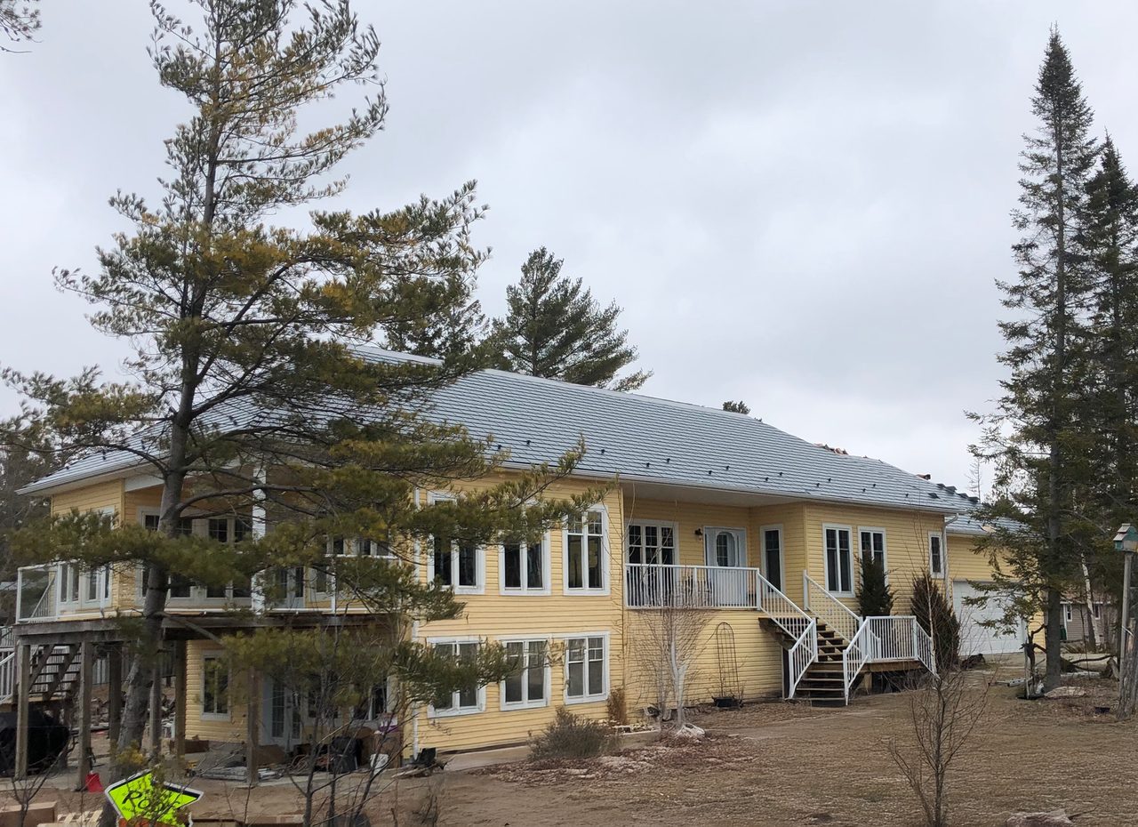 Two-storey yellow cottage with Interlock deep charcoal cedar shingle aluminum hip roof, white railings, and large decks near Wasaga Beach.