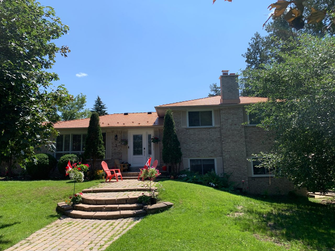 Front elevation of a 1.5-storey brick home in Aurora, ON featuring Interlock Copper Penny cedar shingle metal roofing with a chimney and mature tree surround.