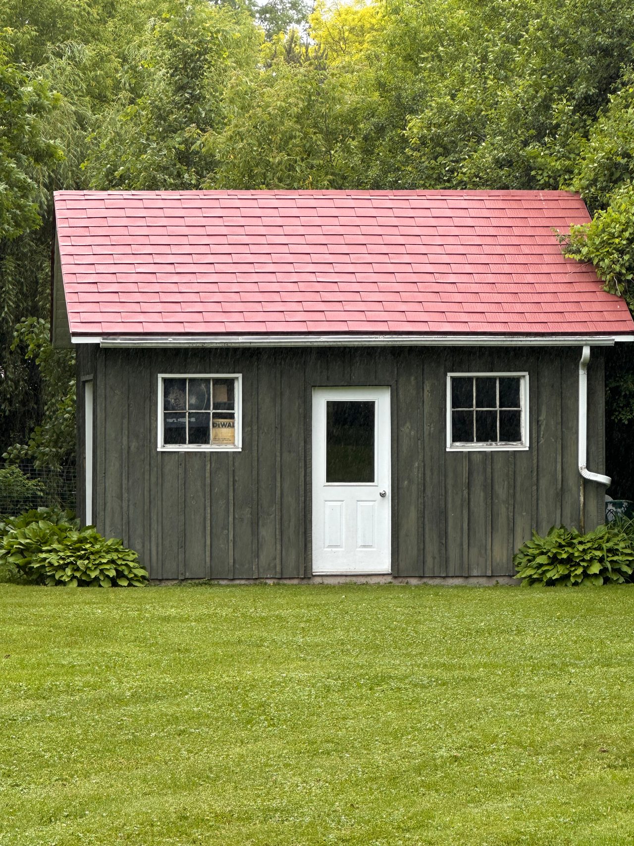 Front elevation of a rustic wood-clad outbuilding with a Tile Red Interlock aluminum shingle gable roof, white door, and twin windows in Almonte, ON.