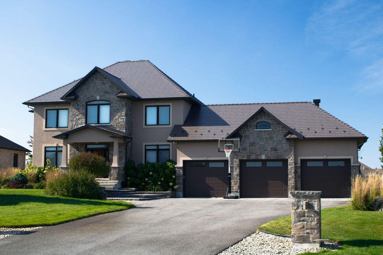 Two-storey stone-and-stucco home in Greely ON with Interlock Tudor Brown cedar shingle metal roofing across a complex multi-hip roof.