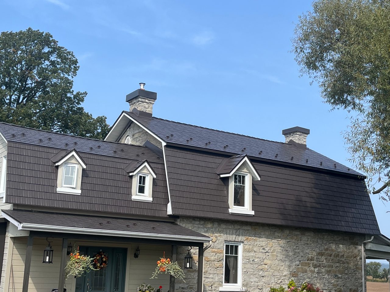 Tudor Brown Interlock aluminum cedar shingle metal roof on a 1.5-storey heritage stone home in Rockland, ON, featuring three dormers and two stone chimneys.