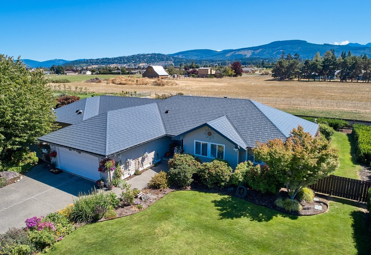 Aerial front view of a ranch-style home in Sequim, WA featuring a complex multi-hip Interlock aluminum slate roof in deep charcoal, with mountain backdrop.