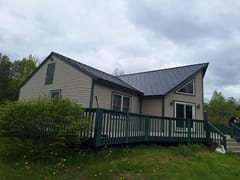 1.5-story rural farmhouse with Interlock deep charcoal aluminum slate metal roof, complex multi-plane gable design, tan vinyl siding, and dark green deck in Stetson, ME.