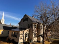 Two-story farmhouse in Russell, MA with Interlock Musket Grey aluminum slate metal roof, steep gable profile, and chimney, framed by bare winter trees.