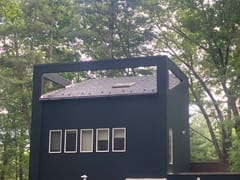 Modern black two-story home with Interlock deep charcoal aluminum slate roof, skylight, and vent pipe visible above a row of horizontal windows, surrounded by forest.