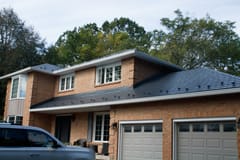Two-storey brick colonial home with deep charcoal Interlock aluminum slate metal roof, snow guards visible, set against a forested suburban backdrop.