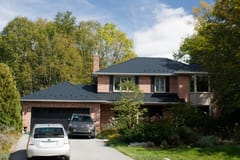 Two-storey brick Colonial home in Kingston, ON with a black Interlock aluminum slate metal roof, chimney, and lush tree-lined suburban setting.
