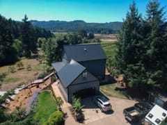 Aerial view of a two-story farmhouse-style home in rural Veneta, OR featuring a black standing seam aluminum metal roof with complex multi-plane geometry.