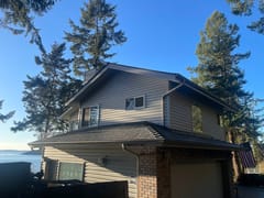 Two-story waterfront home in Anacortes WA with Interlock deep charcoal aluminum slate metal roofing, surrounded by tall evergreens with Puget Sound views.