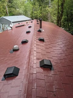 Aerial view of a tile-red Interlock aluminum slate metal roof on a single-family home, featuring multiple roof louvers, a vent pipe, and chimney, surrounded by forest.