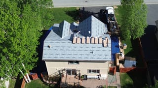 Aerial view of a finished metal roof on a ranch style house in a suburban area with lush greenery and blue sky.
