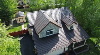 A brown metal roof on a white cottage with brown trim, set amidst lush greenery.