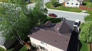 A modern single-family home with a brown metal roof, viewed from above, in a rural or suburban setting with lush greenery and blue sky